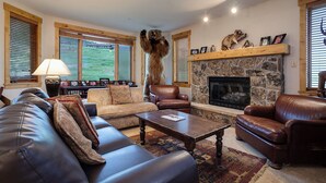 A cozy living room with leather sofas, a stone fireplace, and large windows with blinds.