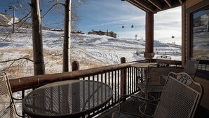 A covered outdoor seating area with metal chairs and a table, overlooking a snowy mountain with ski lifts.