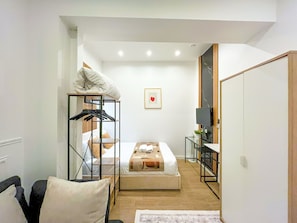 A compact hotel room with a bed, a seating area with a chair and a small table, a wardrobe, and a TV.