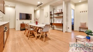 Eine moderne Küche mit Holzschränken, ein Essbereich mit weißem Tisch und Stühlen sowie ein Wohnbereich mit einer Couch und einer Pflanze.