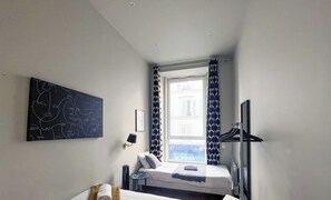 A small, modern hotel room with a single bed, a bedside table, a wall-mounted TV, and a large window with patterned curtains.