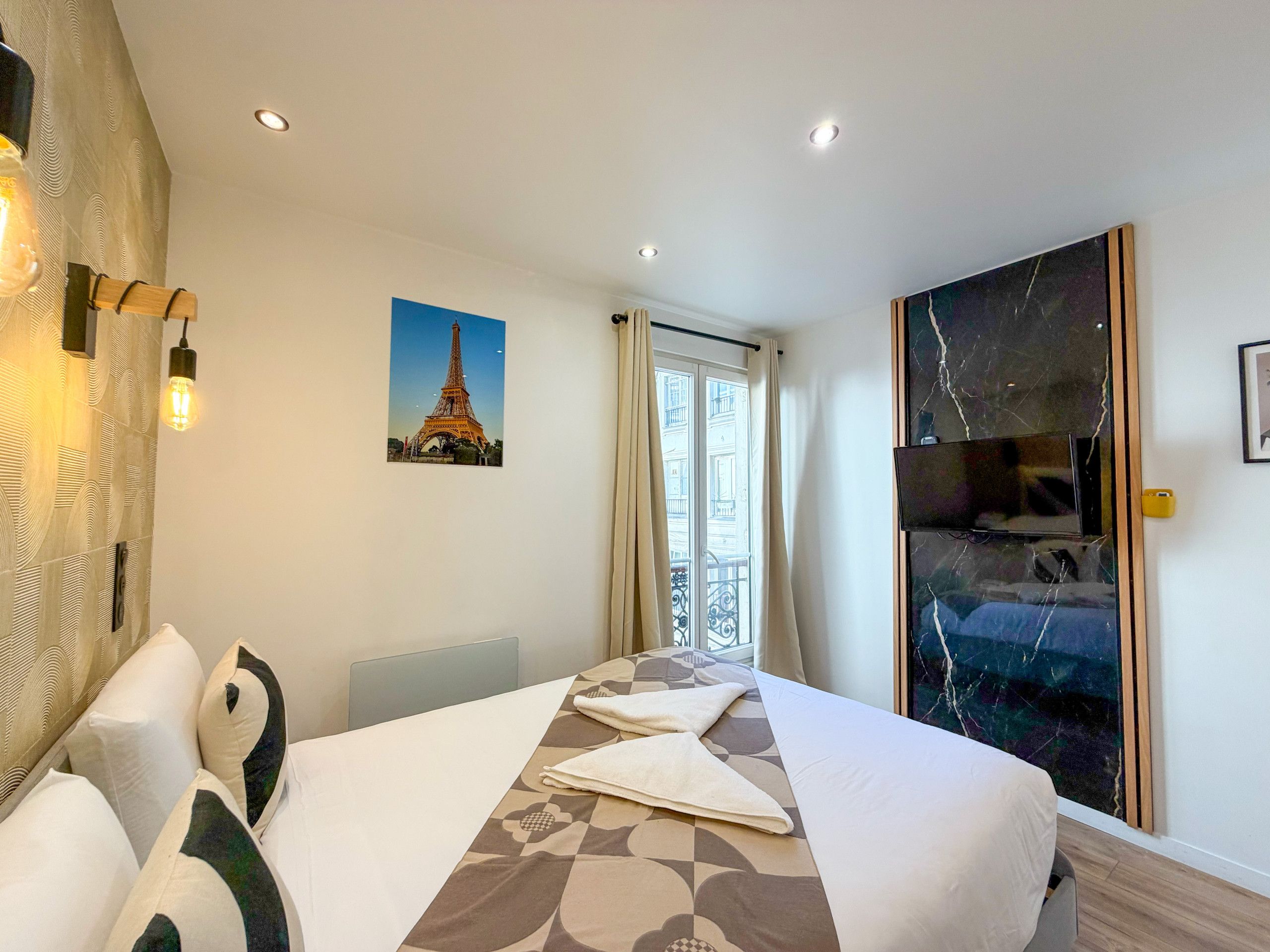 Una habitación de hotel con una cama, un cuadro de la Torre Eiffel, una ventana grande con cortinas y una obra de arte de mármol en la pared.