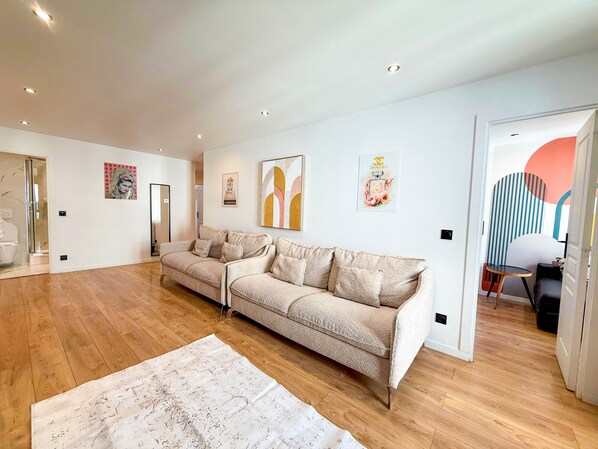 Un vaste espace de vie agrémenté d’un canapé beige, d’un parquet en bois et d’œuvres d’art accrochées aux murs.