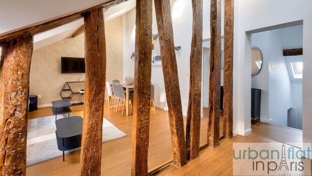 A modern interior with exposed wooden beams, a dining area with a round table and chairs, and a flat-screen TV mounted on the wall. - Urban Flat  117 - Charming 3BDR Apartment in Paris (París)