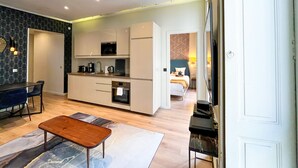 Eine moderne Küche mit weißen Schränken, einer Mikrowelle und einer Kaffeemaschine. Ein Essbereich mit Tisch und Stühlen ist ebenfalls vorhanden. Im Hintergrund ist ein Bett zu sehen.