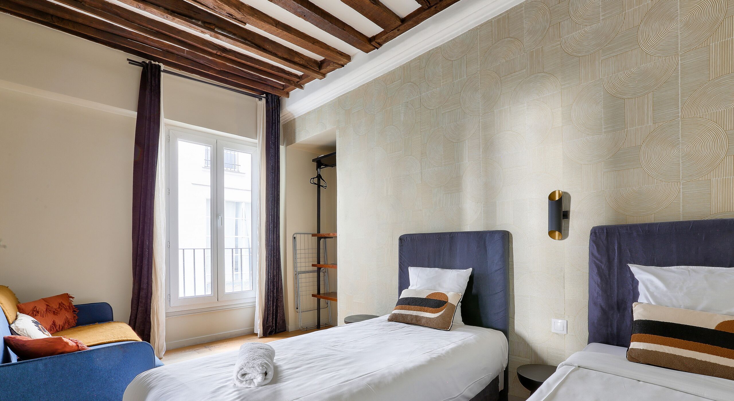 Une chambre d’hôtel avec deux lits, une fenêtre avec des rideaux, un plafond en bois et un papier peint à motifs.
