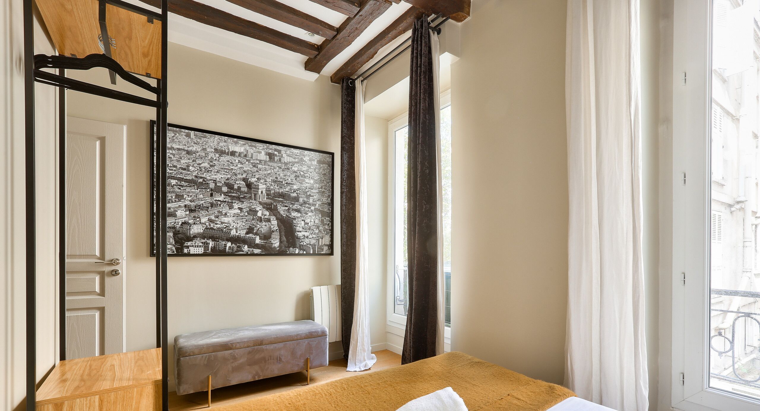 Una camera d'albergo con un letto, una panca, un grande quadro in bianco e nero raffigurante un paesaggio urbano e una finestra con tende.