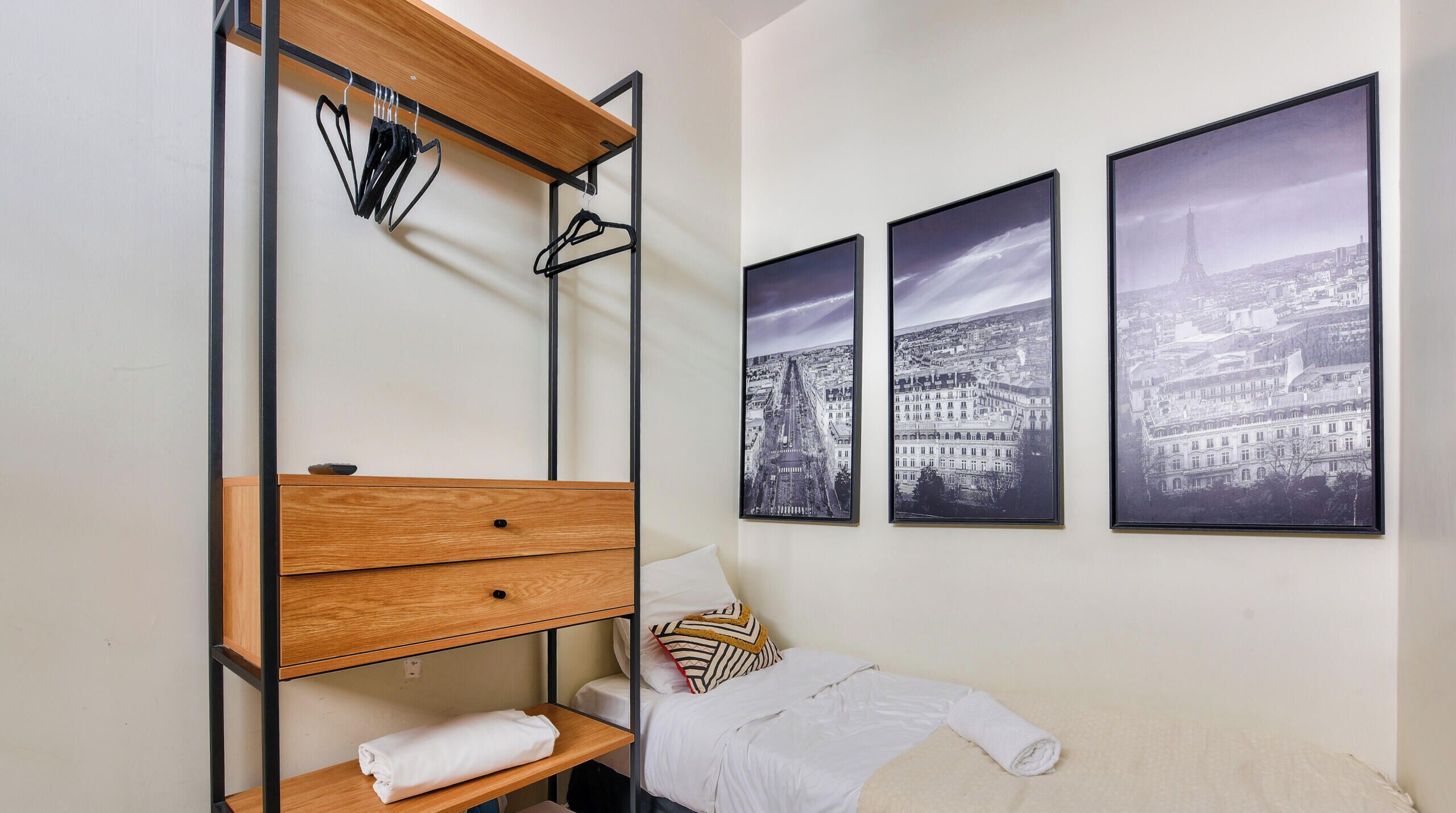 A bedroom with a bed, a wooden dresser, a shelf, and three black and white cityscape photographs on the wall.