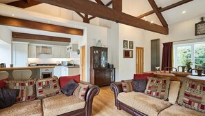 A spacious living area with a fireplace, wooden beams, a kitchen in the background, and a large window with curtains. - Host & Stay | River View Cottage (Barnard Castle)