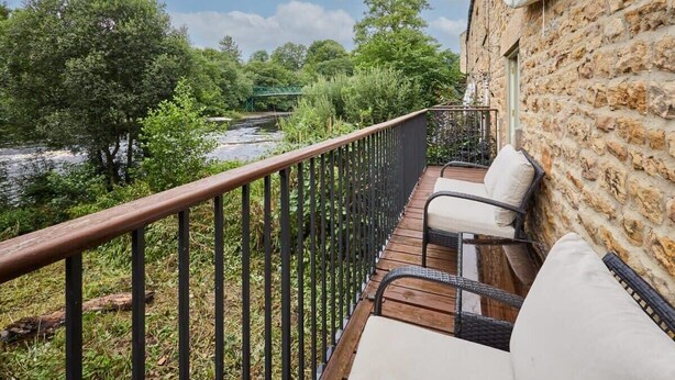 A balcony with wicker furniture and a stone wall, overlooking a river and greenery. - Host & Stay | River View Cottage (Barnard Castle)