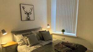 A neatly made bed with a grey blanket and pillows, a bedside lamp, a small plant on the windowsill, and a framed deer picture on the wall. - Deeside Snugs (Aboyne)