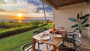 An outdoor dining area with a view of the sunset over the ocean, a table set with plates, cutlery, and a pineapple. - Keauhou Kona Surf & Racquet 1104: Ocean Views, Resort Amenities & Prime Location (Kailua Kona)