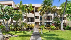 A multi-story building with a central pathway leading to the entrance, surrounded by lush greenery and palm trees. - Keauhou Kona Surf & Racquet 1104: Ocean Views, Resort Amenities & Prime Location (Kailua Kona)