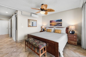 A bedroom with a bed, a bench, a nightstand, a lamp, a ceiling fan, a painting, and a door. - Keauhou Kona Surf & Racquet 1104: Ocean Views, Resort Amenities & Prime Location (Kailua Kona)