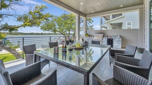 Un espace de restauration extérieur moderne, avec des meubles en osier, une table en verre et une vue sur l’eau et les arbres.