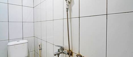 Un baño con paredes de azulejos blancos, inodoro, ducha con cabezal de mano y tubería visible.