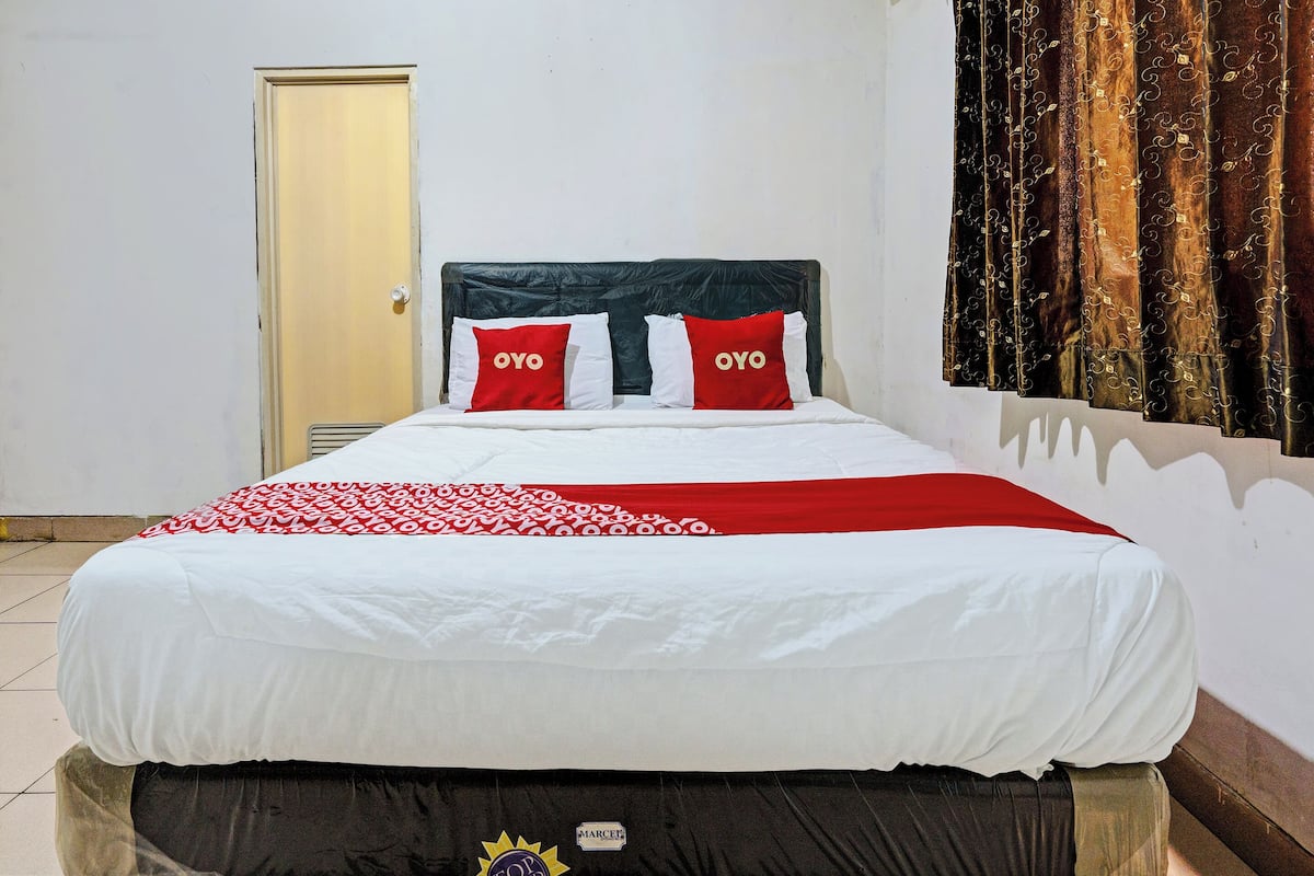 Un lit avec une couvre-lit blanche et rouge, deux oreillers rouges ornés du texte « OYO », une tête de lit sombre et un rideau à motifs de feuilles.