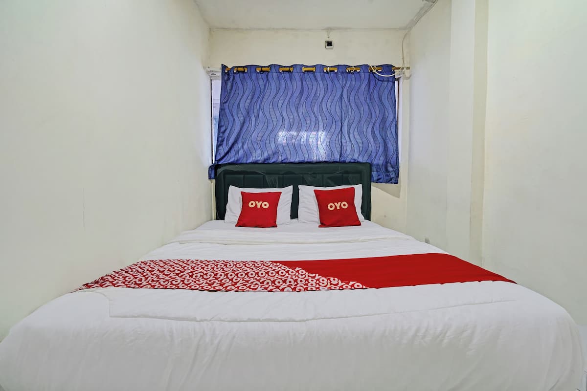 Un lit simple avec une couvre-lit blanche et rouge, deux oreillers rouges ornés du logo « OYO », et un rideau bleu au-dessus.