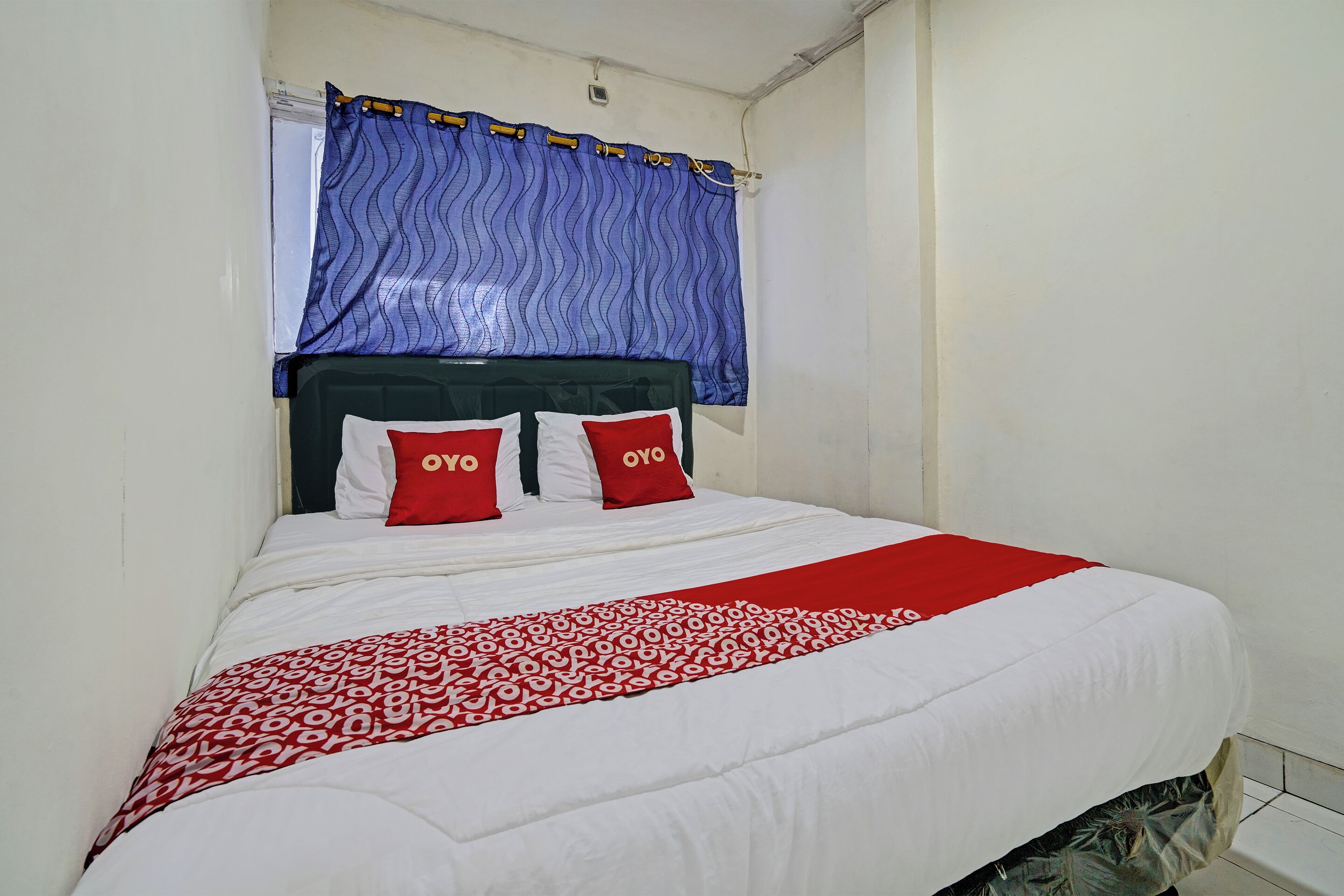 A neatly made bed with a white and red bedspread, two red pillows with "OYO" text, and a blue curtain above.