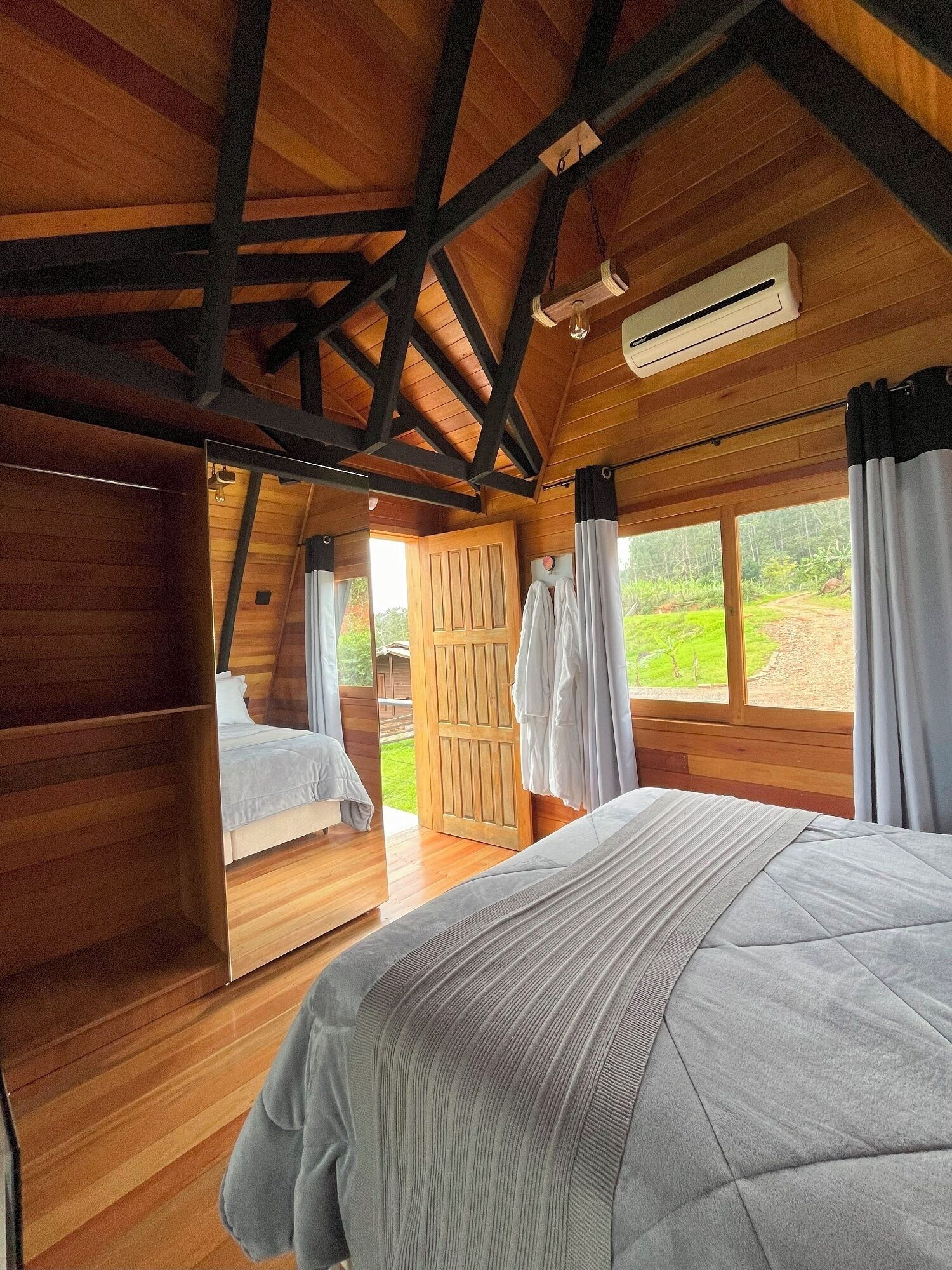 Ein Schlafzimmer mit Holzbalkendecke, einem Bett mit grauer Steudecke und Blick auf eine grüne Landschaft durch ein Fenster.