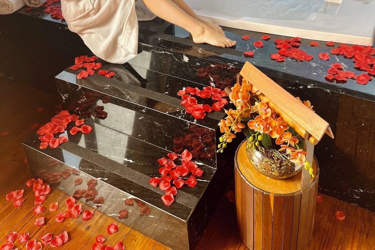 Una persona vestida con bata, sentada junto a un jacuzzi con pétalos de rosa y un globo con forma de corazón, contemplando un paisaje pintoresco.
