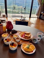 A breakfast table with various dishes, including toast, fruit, and an omelette, set against a backdrop of a scenic view through large windows.