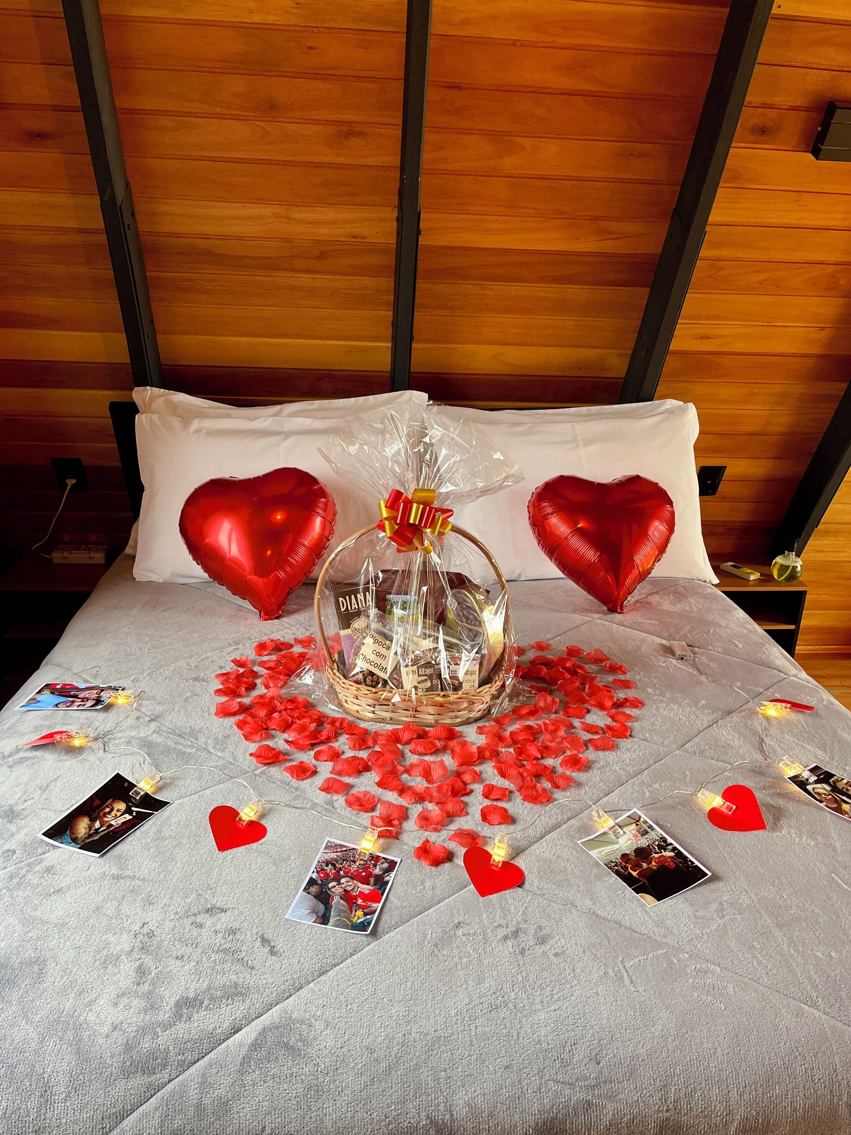 A bed with a white bedspread, two heart-shaped pillows, a basket with a bow, and scattered red heart-shaped decorations.