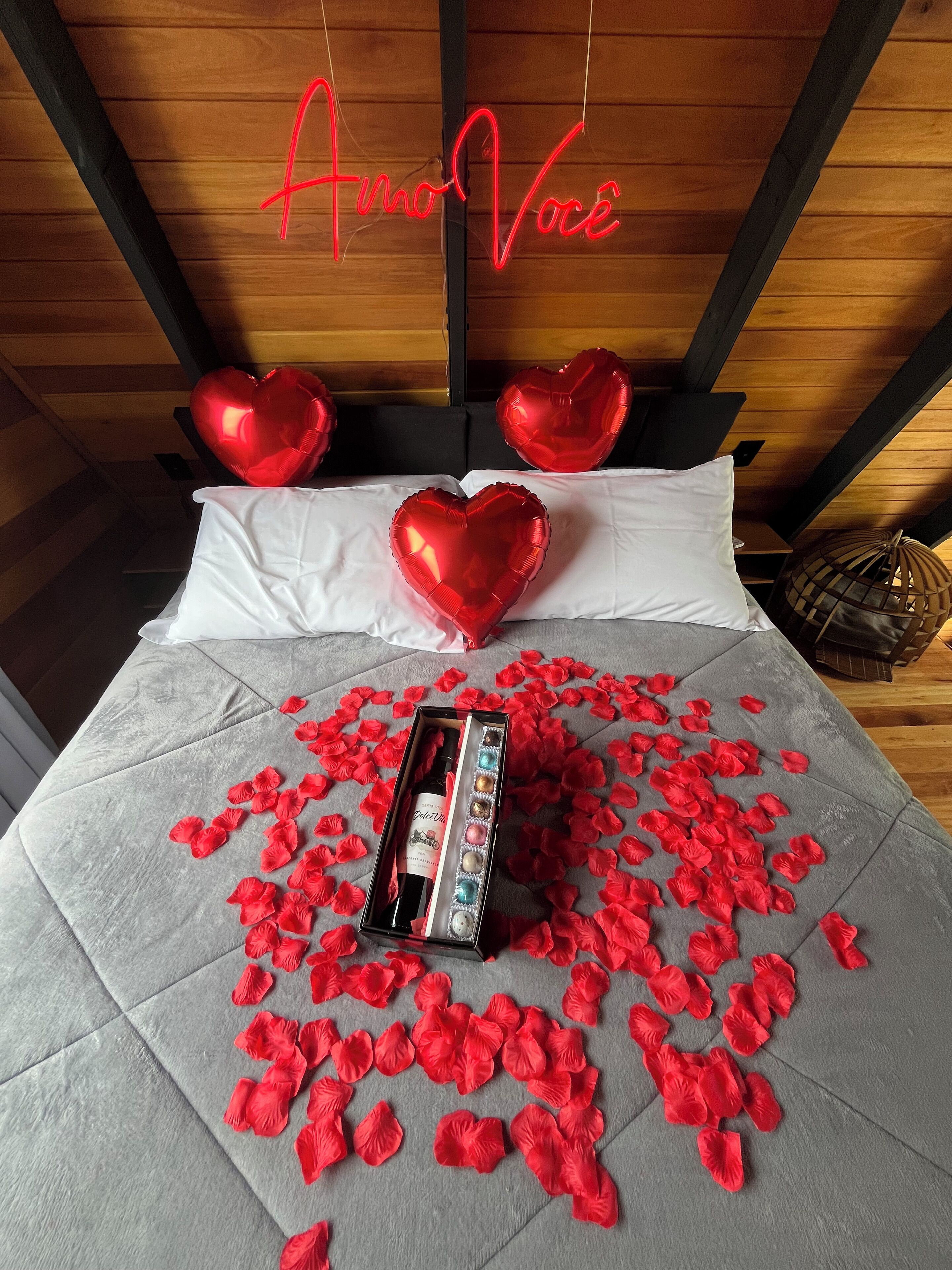 Una cama con globos con forma de corazón rojo, una botella y pétalos de rosa sobre una colcha gris.