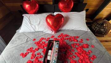 Una cama con globos con forma de corazón rojo, una botella y pétalos de rosa sobre una colcha gris.