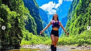 Una persona de pie sobre una roca junto a un río, con abundante vegetación y montañas al fondo.