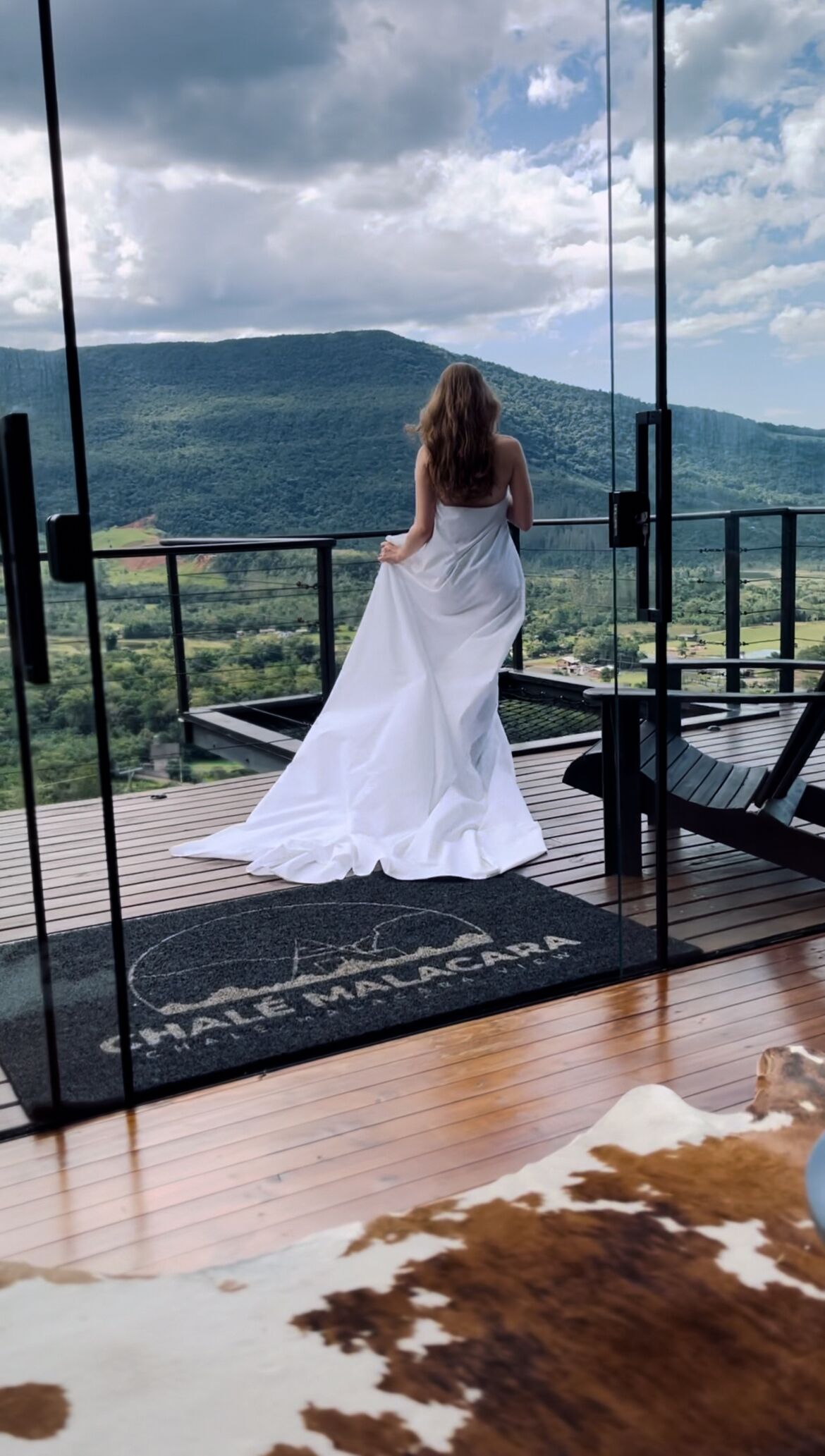 A person in a white dress standing on a balcony with a view of a mountainous landscape.
