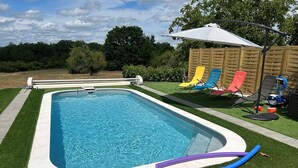 A rectangular swimming pool with a white edge, surrounded by artificial grass. There are colorful lounge chairs and a white umbrella in the background.