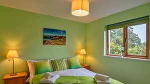 A neatly made bed with green and white bedding, two bedside lamps, a painting on the wall, and a window with a view of greenery.