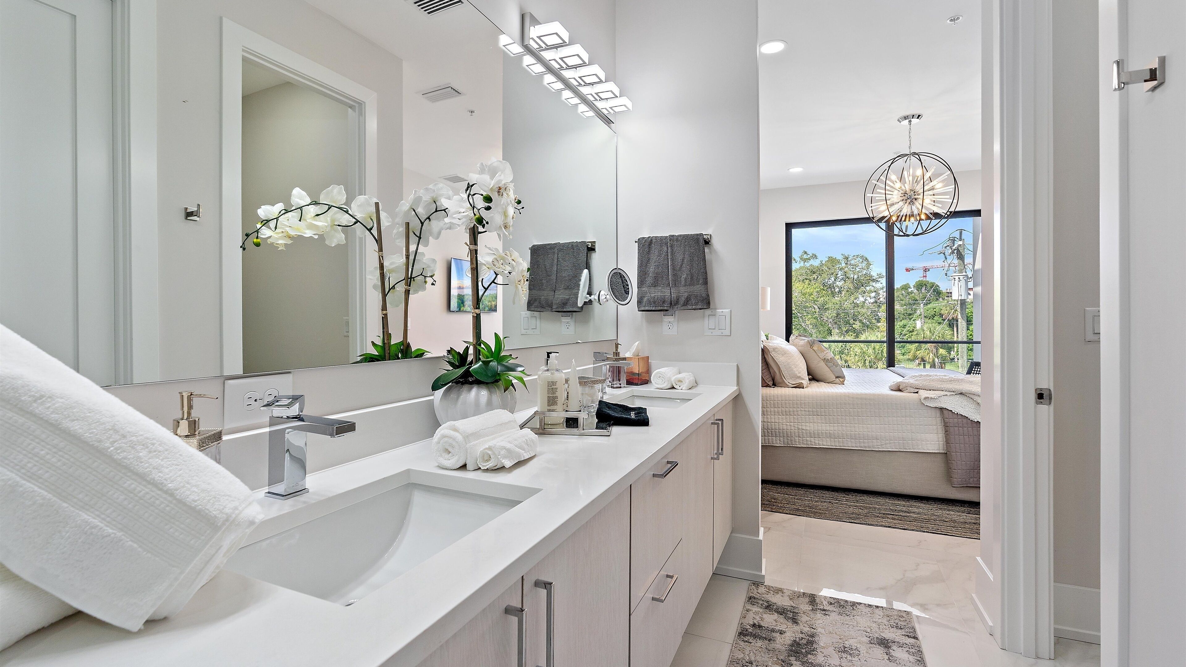 Une salle de bain moderne avec un grand miroir, un lavabo blanc et une vue sur une chambre à coucher avec un lit et un balcon.