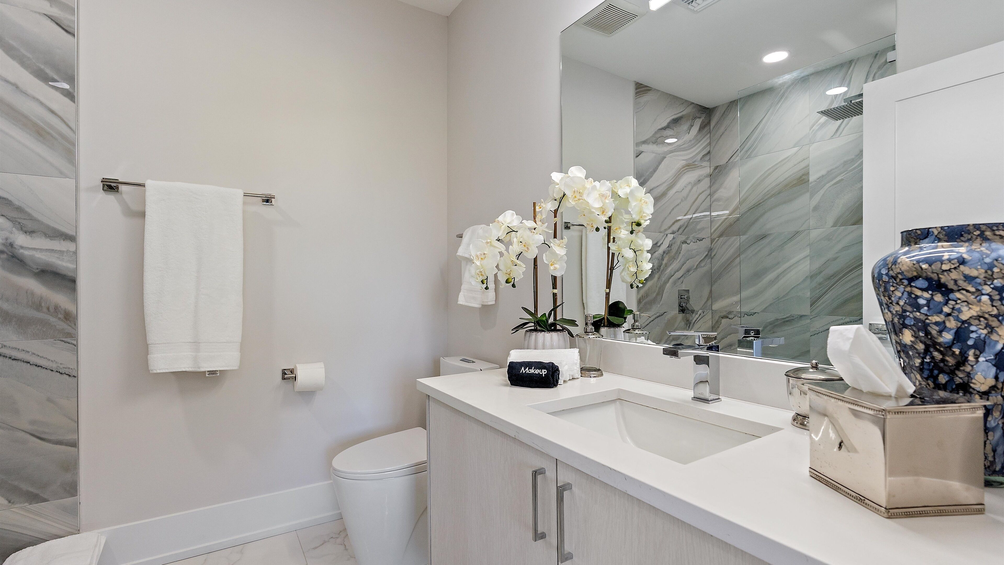 A modern bathroom with a marble backsplash, a large mirror, a white sink, and a white toilet.