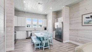A modern kitchen with white cabinetry, a stainless steel refrigerator, and a dining area with a blue table and chairs. - Birds Eye View Sp163 3 Bedroom Home by RedAwning (Rockport)