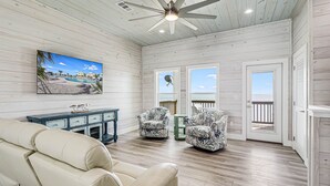A spacious room with a wooden ceiling, a ceiling fan, a large window with a view of the beach, two patterned armchairs, a beige sofa, and a small table. - Birds Eye View Sp163 3 Bedroom Home by RedAwning (Rockport)