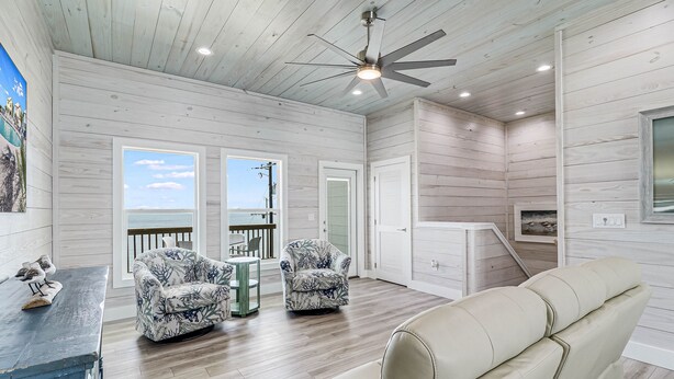 A spacious room with a ceiling fan, wooden walls, a large window, a balcony, and a seating area with a sofa and chairs. - Birds Eye View Sp163 3 Bedroom Home by RedAwning (Rockport)