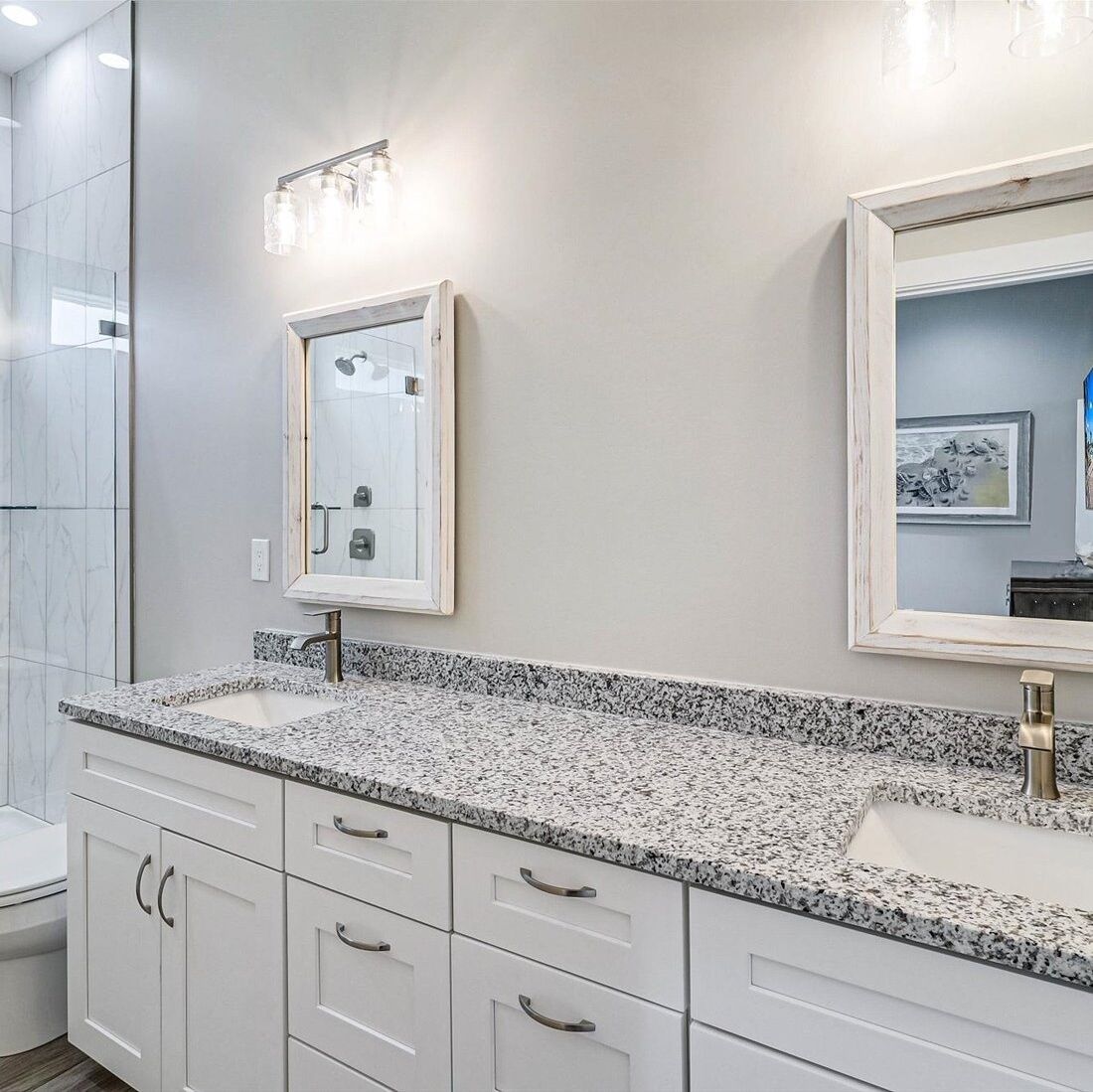 A bathroom with a granite countertop, white cabinets, a framed mirror, and a shower enclosure.