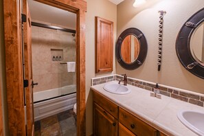 Une salle de bain avec deux lavabos, un meuble-lavabo en bois, une douche et un miroir suspendu à une chaîne.