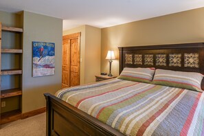 Une chambre avec un lit en bois, une couverture à rayures colorées, une table de chevet en bois avec une lampe et une affiche accrochée au mur.
