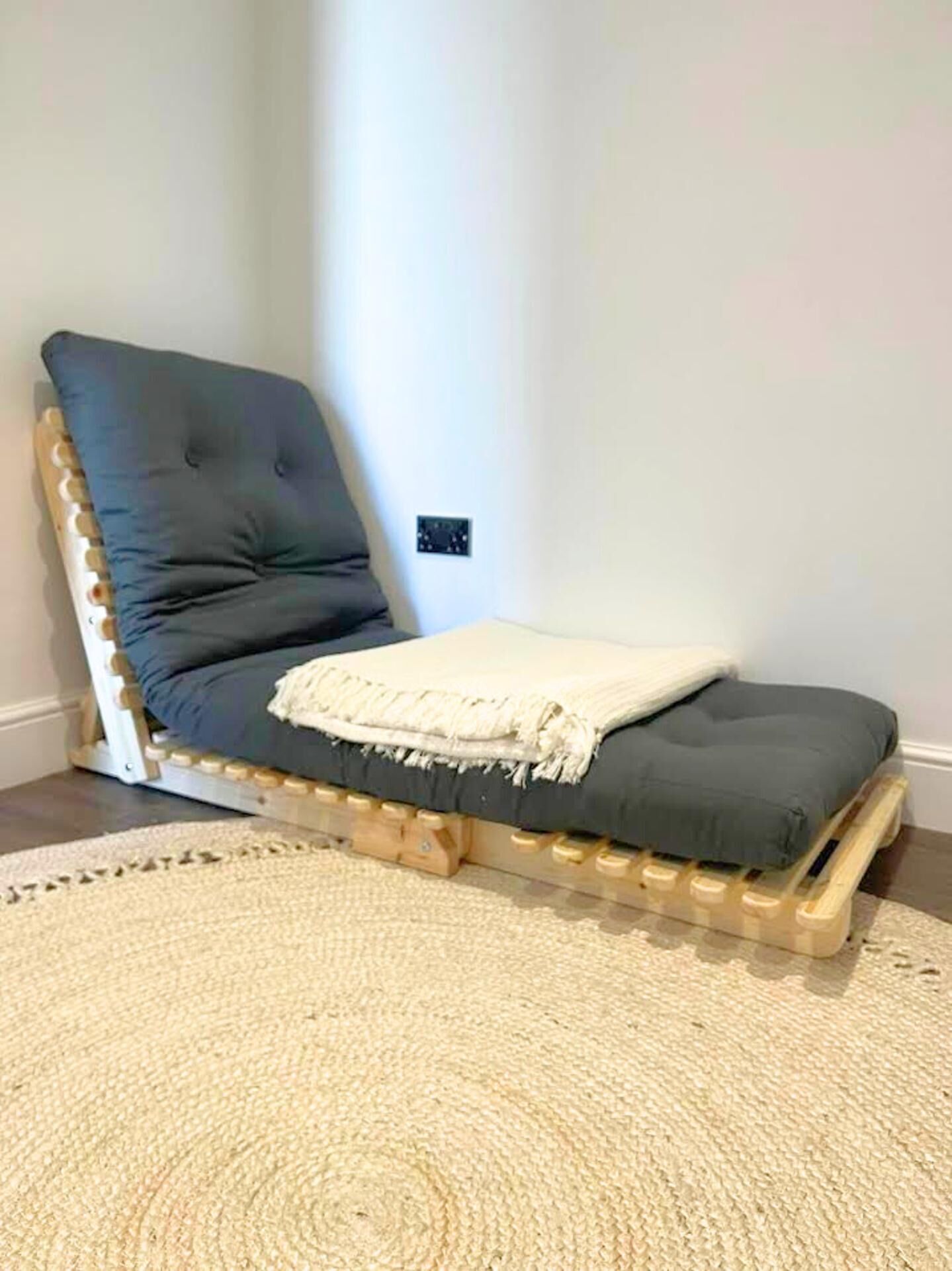 Un lit simple avec un matelas gris et une couverture blanche, placé contre un mur blanc près d’une prise de courant.