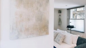 Ein Zimmer mit einem großen, abstrakten Gemälde an der Wand, einem weißen Sofa mit gemusterten Kissen, einem Beistelltisch und einem Fenster mit Jalousien.