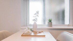 Ein schlicht eingerichtetes Zimmer mit großem Fenster, weißem Tisch und zwei Topfpflanzen.