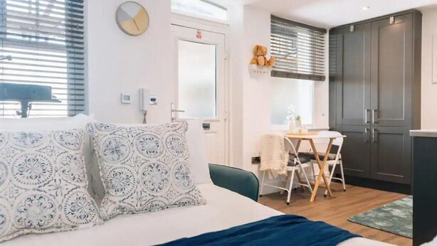 Ein Schlafzimmer mit Bett, Kissen, Nachttisch, Fenster mit Jalousien, einer Wanduhr und einer Tür.
