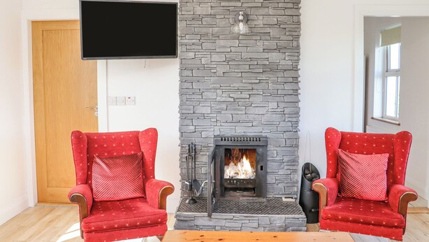 A living room with a stone fireplace, two red armchairs, a flat-screen TV, and a wooden coffee table. - An Teachain (Adamstown)