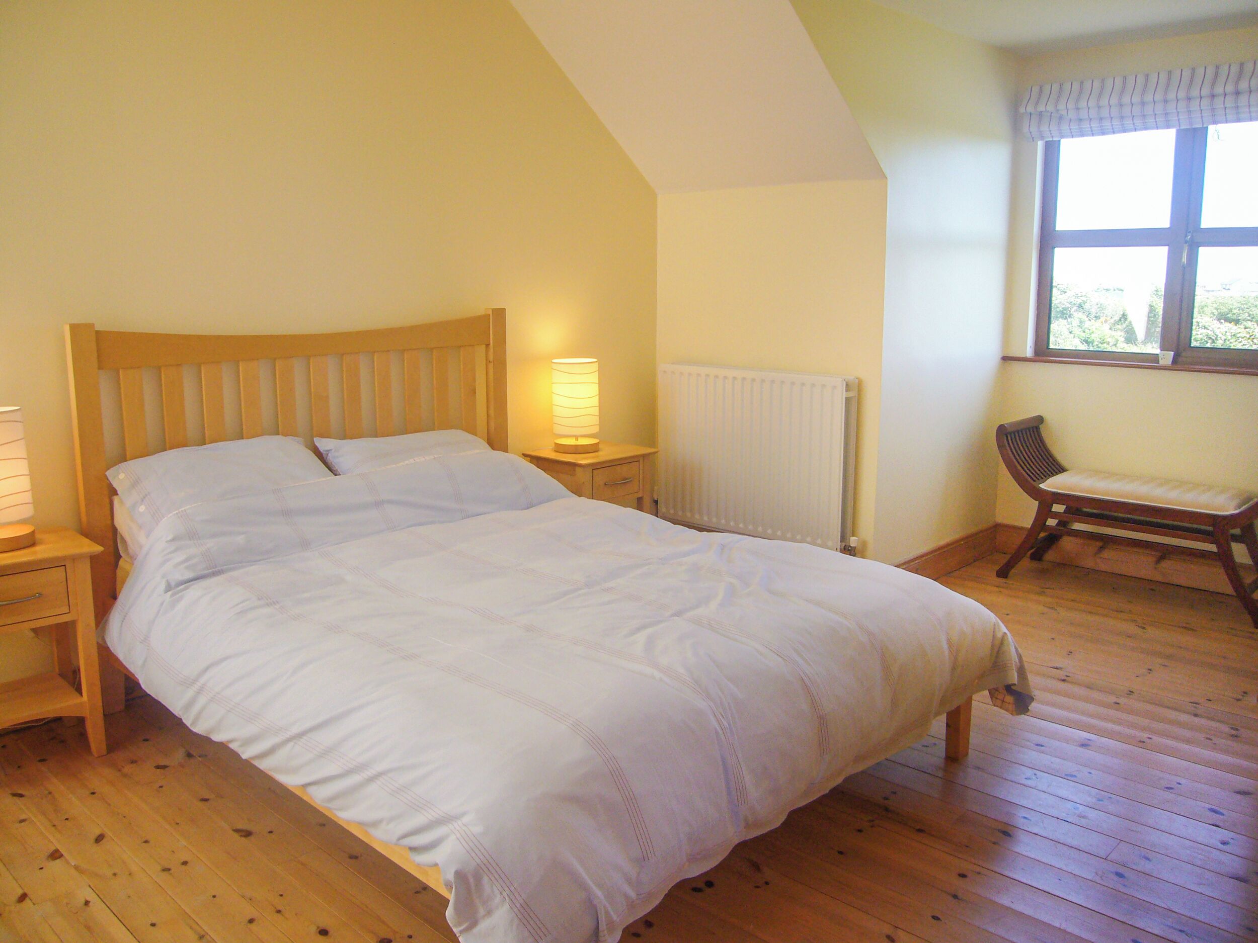 A bedroom with a large bed, wooden headboard, bedside tables with lamps, a radiator, a window with curtains, and a wooden bench.