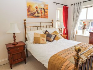 Une chambre à coucher avec un lit, une table de chevet, une lampe, un tableau, une fenêtre avec des rideaux et une vue sur l’extérieur.