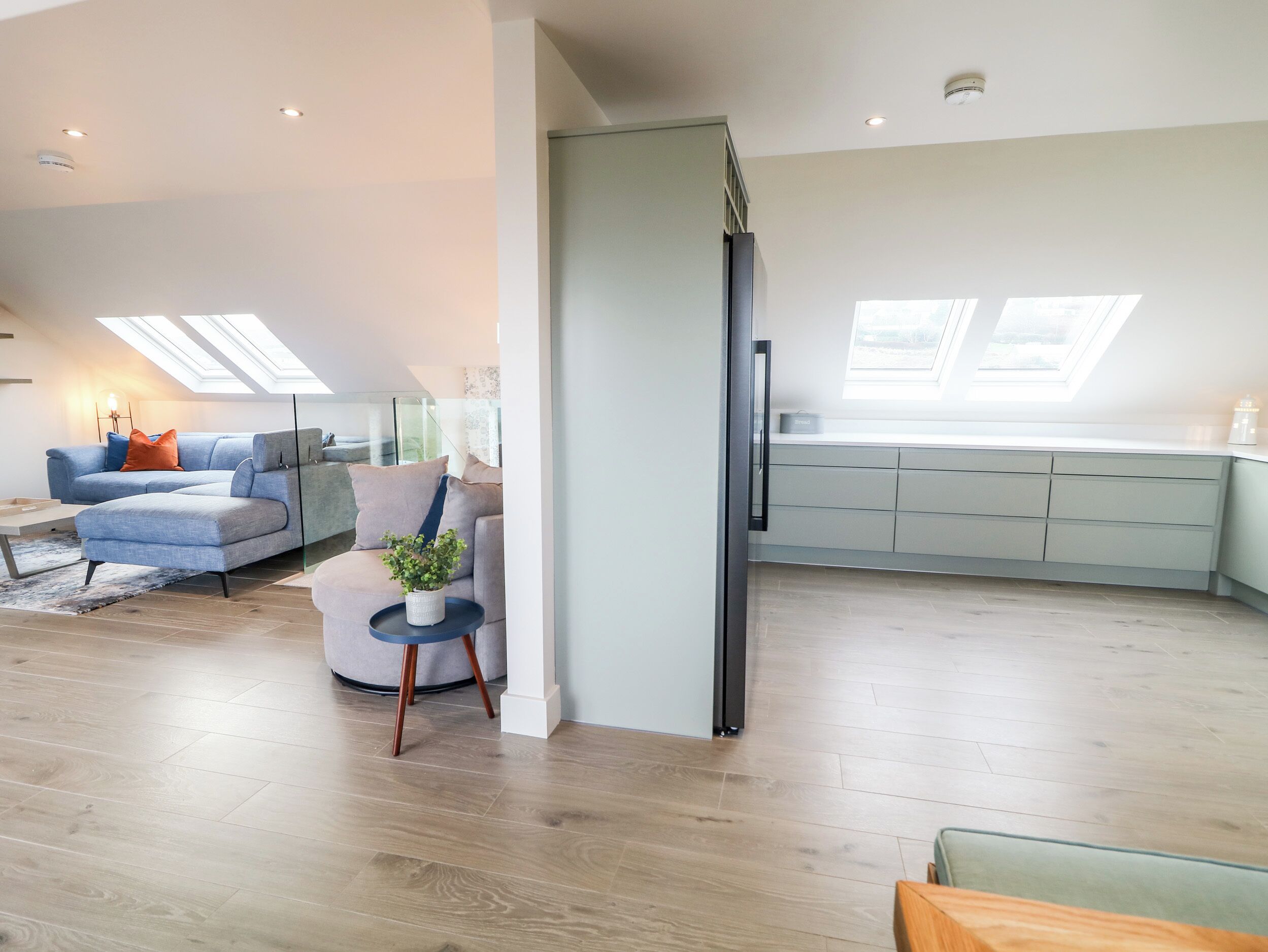 A modern living room with a sectional sofa, a small round table with a plant, and a kitchen area with cabinets and a countertop.