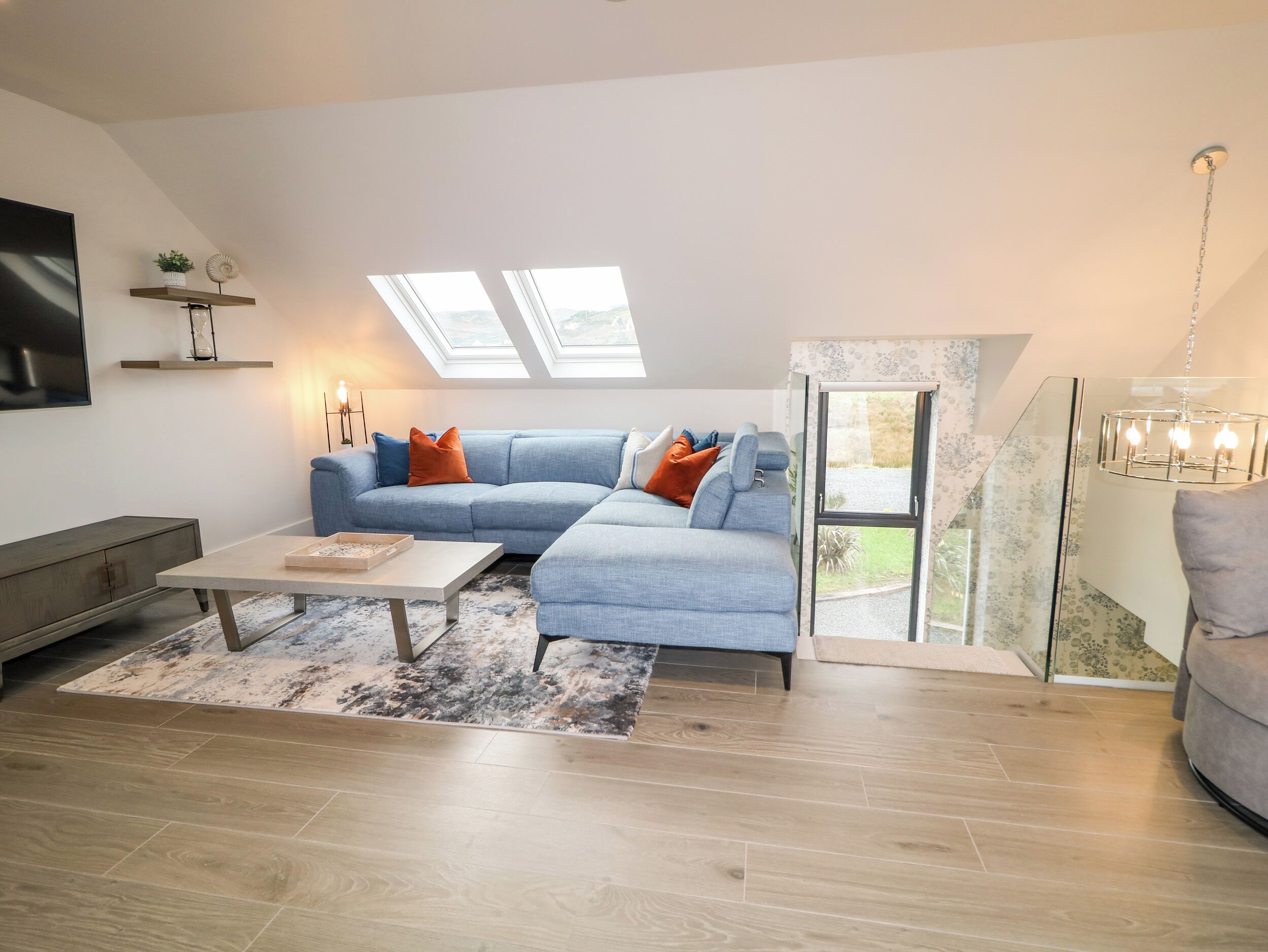 Een moderne woonkamer met een hoekbank, een salontafel en een televisie. Er zijn dakramen en uitzicht op de omgeving.
